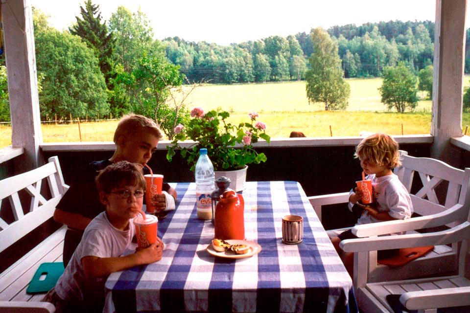 Tre barn som sitter på en veranda och dricker ur röda pappkoppar, idyllisk utsikt